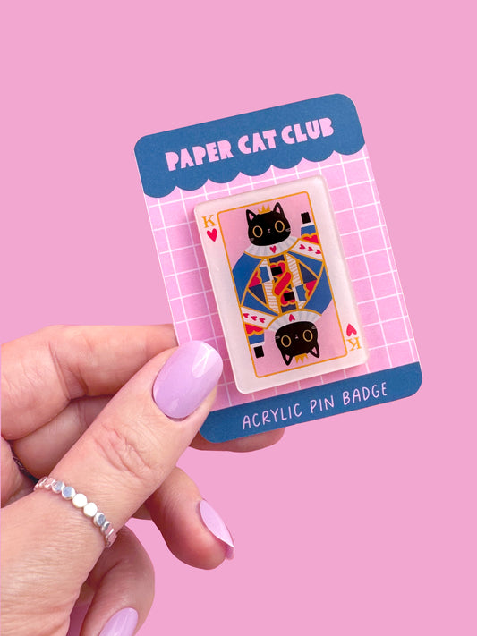 Close-up of a King of Hearts Cat lapel pin on a backing card . Made of acrylic with a pink heart clutch, showing a cat with a crown on a playing card background, a cute cat pin for gifts and collections.