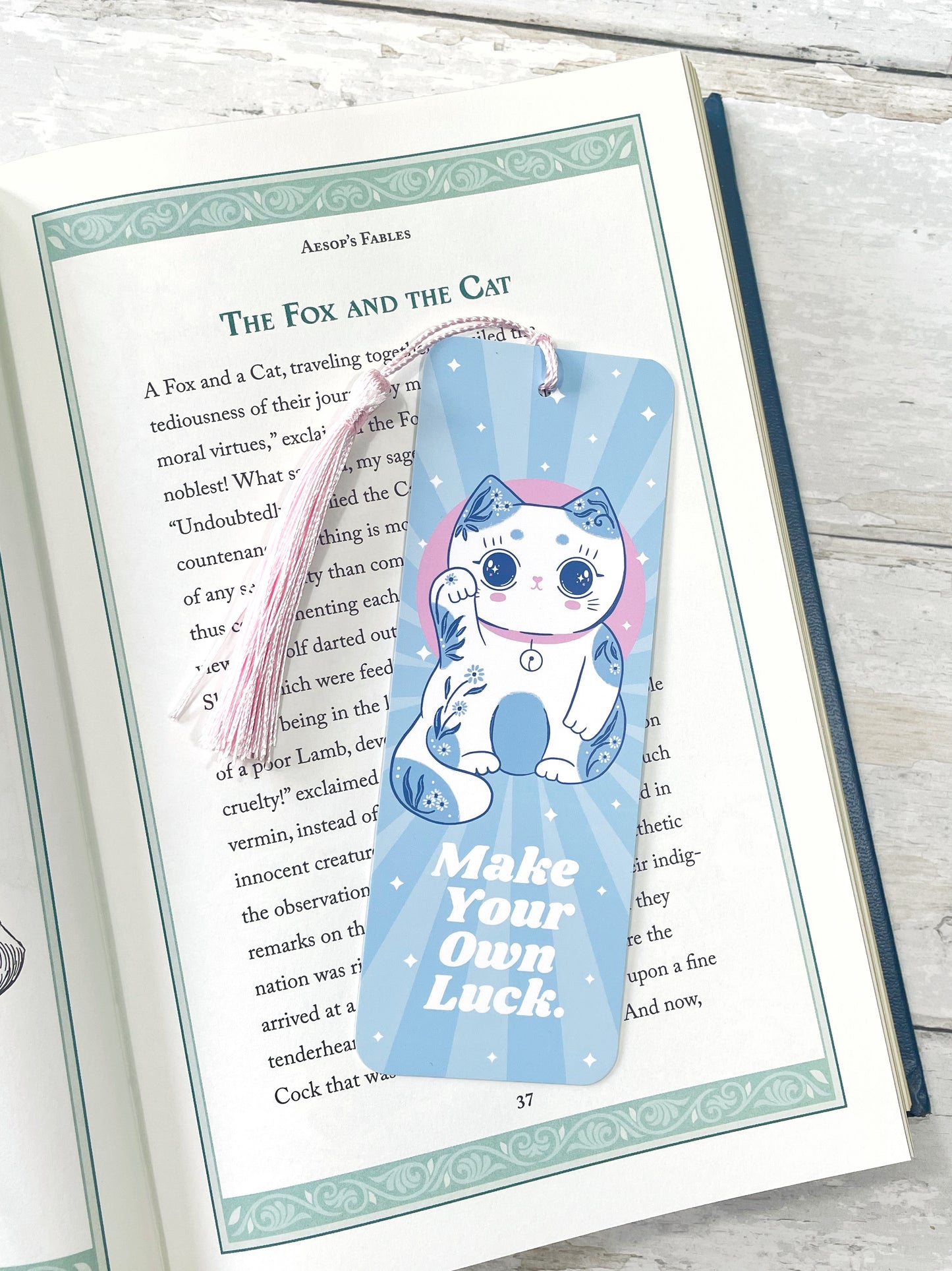 Light blue lucky cat bookmark with pink tassel placed in an open book, featuring the phrase 'Make Your Own Luck,' perfect for kawaii style lovers and book lovers